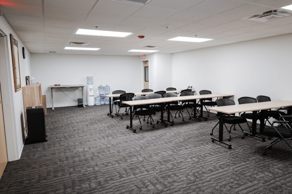 Horizontal image of a open room with 3 tables, lined up with chairs pushed up to them, facing the same direction like a classroom