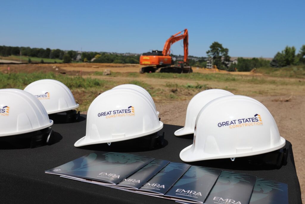 Great States Construction hard hats and Emra Sioux Falls brochures at the groundbreaking, with an excavator on site